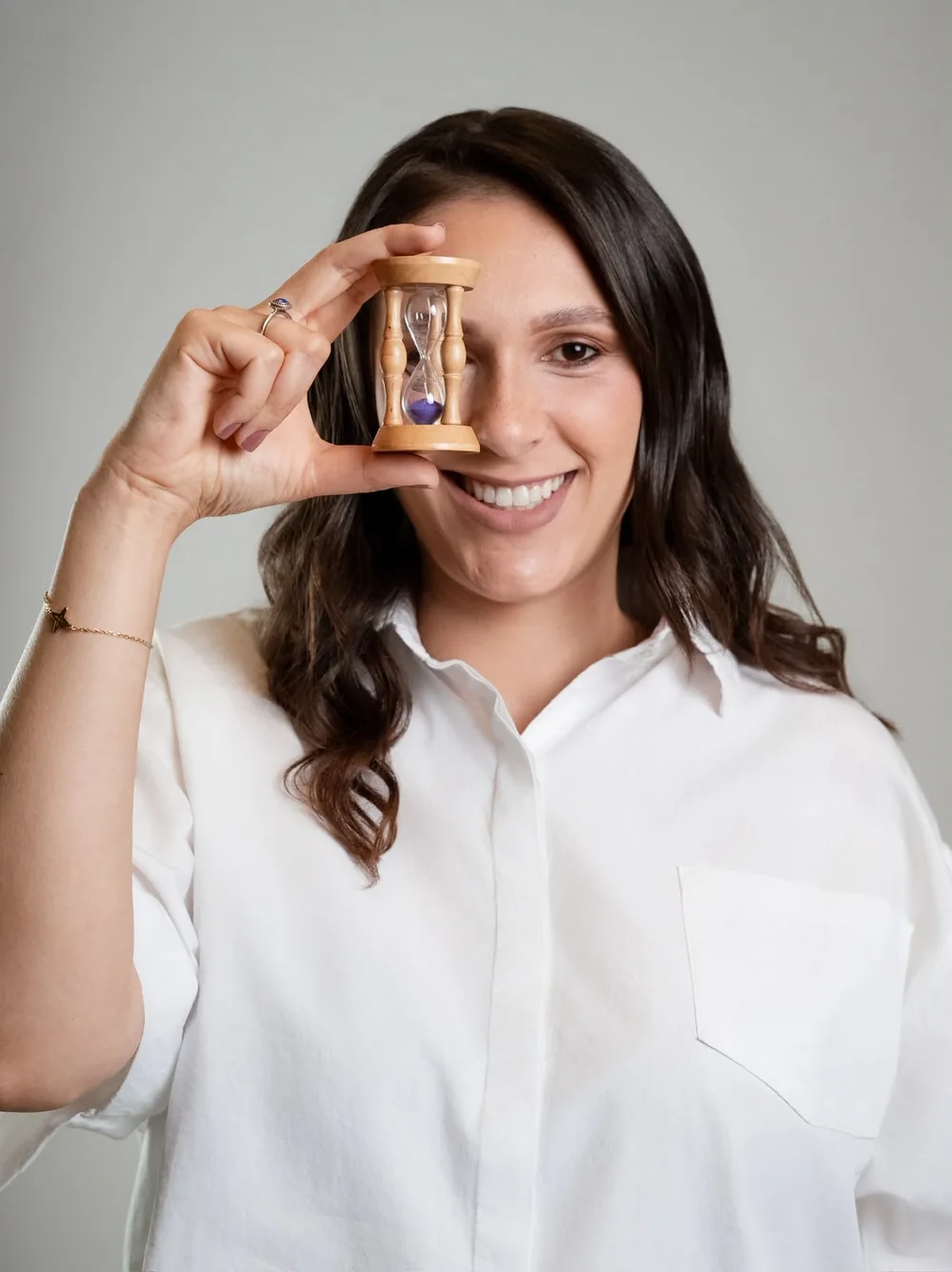 Carla Parreira sorrindo e segurando uma pequena ampulheta de madeira diante do rosto, simbolizando o tempo dedicado a si